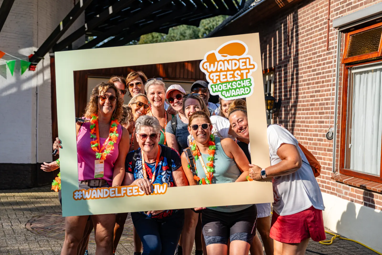 Vrolijke wandelaars met bloemenkransen poseren in een grote fotolijst van het Wandel Feest